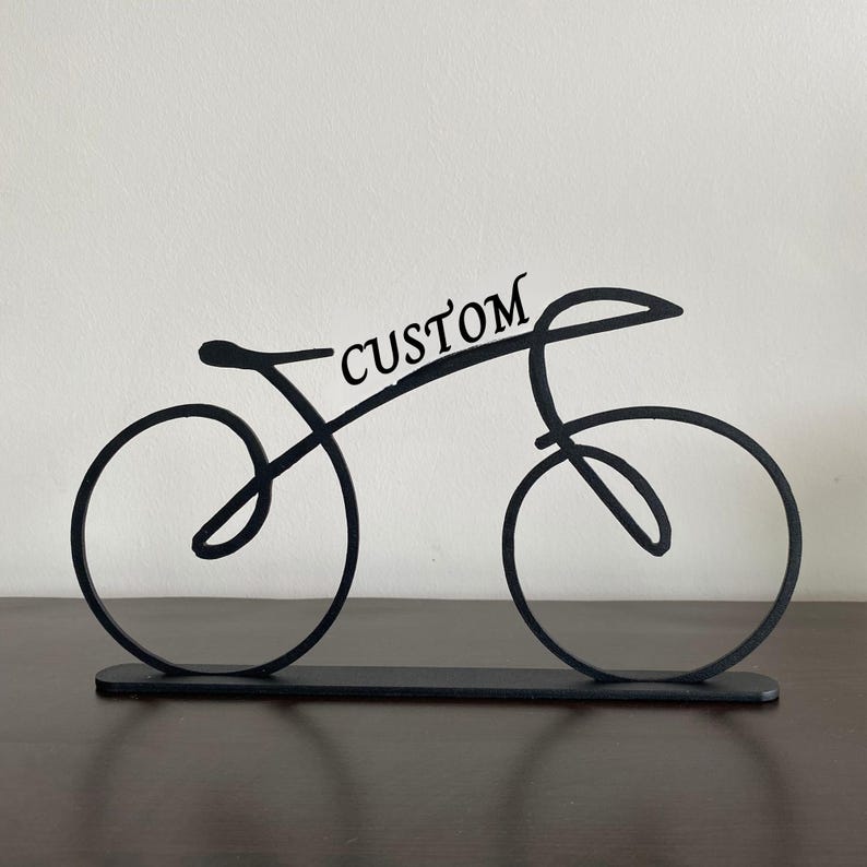 May include: Black metal bicycle sculpture with the word "CUSTOM" in script. The bicycle is a minimalist design with two circular wheels and a curved frame, mounted on a rectangular base.