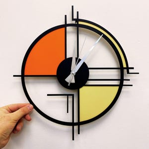 Puede incluir: Un reloj de pared moderno con marco negro y diseño geométrico. La esfera del reloj está dividida en cuatro secciones, dos de las cuales son naranja y amarillo. Las manecillas del reloj son plateadas.