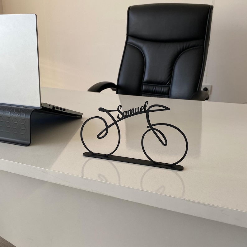 May include: A black metal bicycle ornament with the name "Samuel" in cursive. The desk accessory is on a white desk, with a laptop and black office chair in the background.