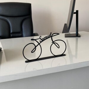May include: Black metal bicycle desk ornament with the name "Samuel" in cursive. The bicycle silhouette has two wheels and a frame, on a flat base. The ornament is on a white desk, with a black office chair and computer monitor in the background.