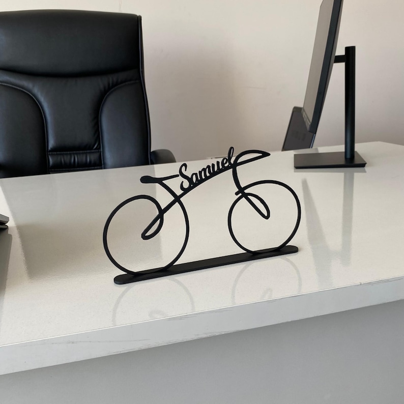 May include: Black metal bicycle desk ornament with the name "Samuel" in cursive. The bicycle silhouette has two wheels and a frame, on a flat base. The ornament is on a white desk, with a black office chair and computer monitor in the background.