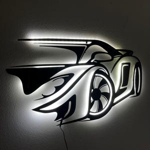 Pode incluir: Uma obra de arte de parede iluminada a preto e branco que retrata um carro desportivo estilizado. O design do carro apresenta iluminação LED branca brilhante que contorna a carroçaria, as rodas e o spoiler. A obra de arte é montada numa parede cinzenta.