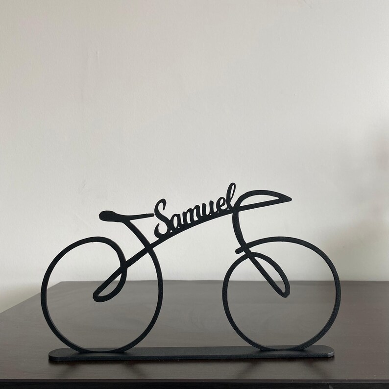 May include: Black metal bicycle sculpture with the name "Samuel" in cursive script. The bicycle has a minimalist design with circular wheels and a sleek frame. It is displayed on a dark surface against a white background.