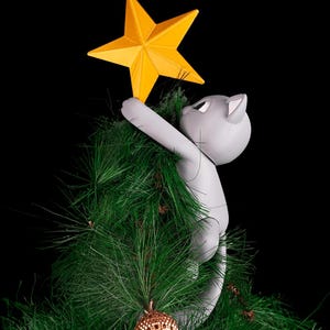 May include: A gray cat-shaped Christmas tree topper reaching for a bright yellow star. The cat is climbing a green artificial Christmas tree, with a gold ornament visible. The background is black.
