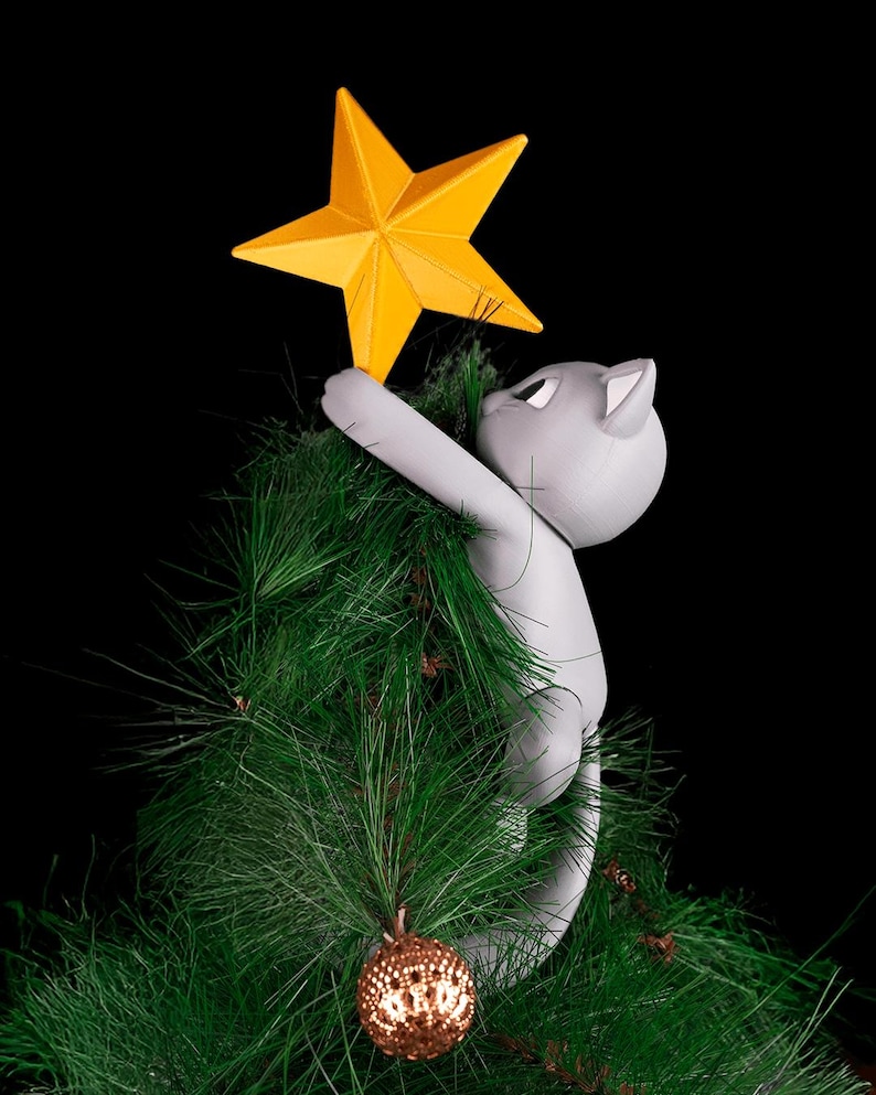 May include: A gray cat-shaped Christmas tree topper reaching for a bright yellow star. The cat is climbing a green artificial Christmas tree, with a gold ornament visible. The background is black.