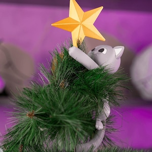 May include: A Christmas tree topper featuring a gray cat reaching for a gold star. The cat is positioned on a green artificial tree, with a purple background. The star is a five-pointed design.