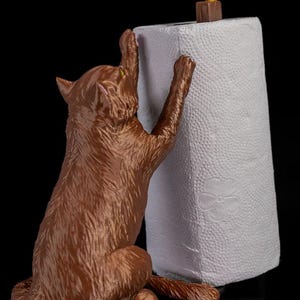 May include: A brown cat figurine holding a white paper towel roll. The cat is reaching up with its paws and has a playful expression on its face. A small yellow bird figurine is perched on top of the paper towel roll.
