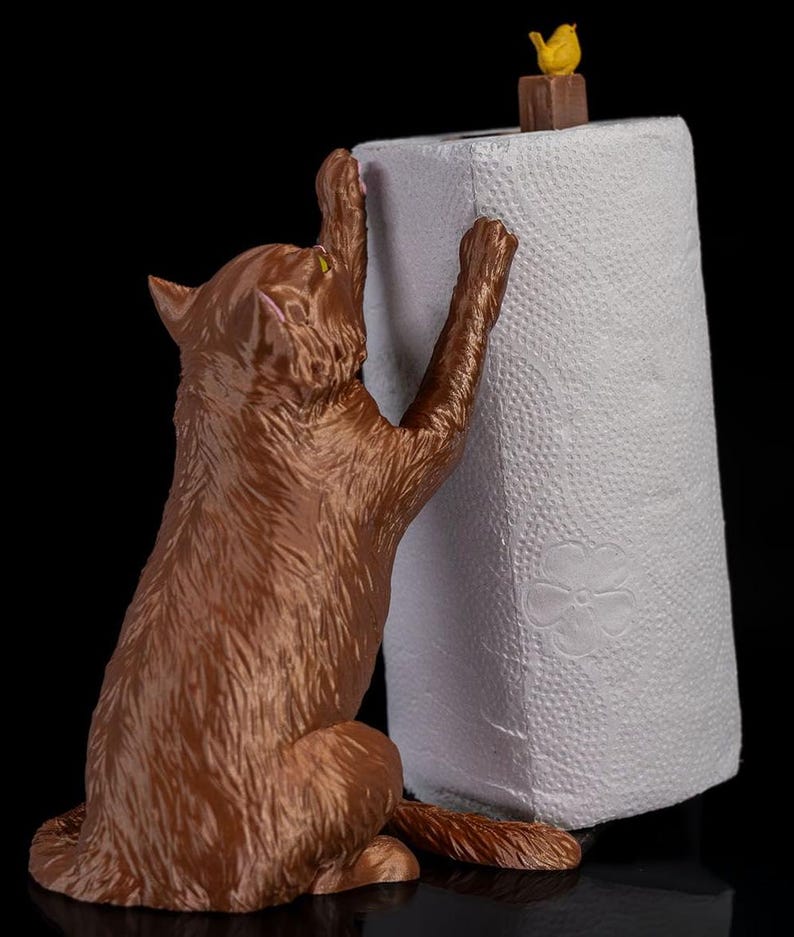 May include: A brown cat figurine holding a white paper towel roll. The cat is reaching up with its paws and has a playful expression on its face. A small yellow bird figurine is perched on top of the paper towel roll.