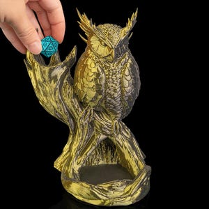 Könnte beinhalten: Goldfarbene Eulenfigur auf einem Ast, mit einem kleinen, türkisblauen, vielseitigen Würfel in der Nähe. Die Eule hat detaillierte Federstrukturen und ist vor einem schwarzen Hintergrund platziert. Die Basis des Astes bildet eine kleine Schale.