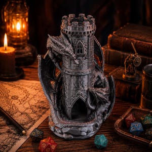 May include: A dark grey, cylindrical dice tower featuring a sculpted dragon wrapped around the exterior. The tower has a crenellated top, a pointed arched doorway at the base, and a small arched window. The surface has a weathered, stone-like texture.