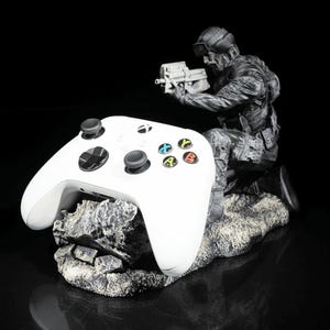Op de afbeelding: Een witte Xbox-controller staat op een rotsachtige basis, ondersteund door een grijs beeldje van een soldaat met een geweer.