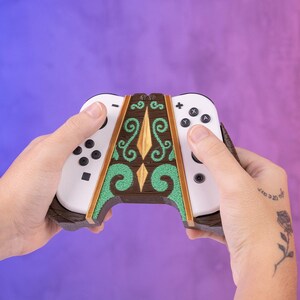 Può includere: Un controller per videogiochi bianco e marrone con un design decorativo in legno. Il controller presenta accenti verdi e dorati, con pulsanti e joystick neri. Il design include un diamante centrale e motivi a spirale.