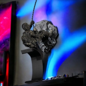 May include: A bronze-colored sculpture of a figure climbing a rock, suspended by a chain. The figure is detailed with textured clothing and gear. The sculpture is mounted on a black, curved base. The background has blue and red lighting.