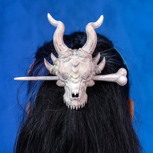 May include: A white dragon skull hair clip with a bone-like design. The clip is attached to a wooden stick.