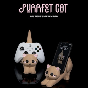 Può includere: Un supporto per telefono e controller di gioco a forma di gatto marrone con occhi rosa e naso bianco. Il supporto è etichettato "PURRFect CAT MULTIPURPOSE HOLDER".