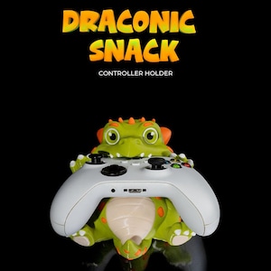 Könnte beinhalten: Ein grüner und orangefarbener drachenförmiger Controller-Halter mit einem weißen Xbox-Controller im Inneren. Der Halter trägt die Aufschrift "Draconic Snack" und "Controller Holder".