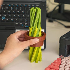 Puede incluir: Una planta de cactus verde impresa en 3D en una maceta marrón. El cactus está hecho de múltiples capas de plástico verde y tiene forma de espiral. La maceta está hecha de plástico marrón y tiene una superficie texturizada.