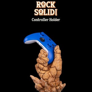 May include: A blue video game controller is held by a rock-like hand sculpture. The sculpture is brown and textured, and the base is made of rock-like crystals. The text "ROCK SOLID! Controller Holder" is above the sculpture.