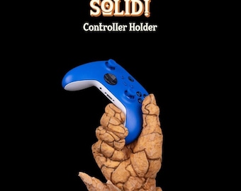 Puede incluir: Un mando de videojuegos azul está sostenido por una escultura de mano de roca. La escultura es marrón y texturizada, y la base está hecha de cristales que parecen rocas. El texto "ROCK SOLID! Controller Holder" está por encima de la escultura.