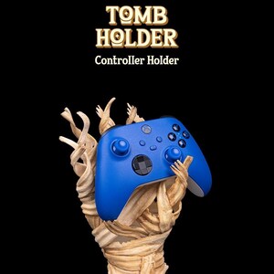 May include: A blue Xbox controller is held by a wooden stand shaped like a mummy. The stand is called "Tomb Holder" and is a controller holder.