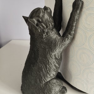May include: Black cat figurine holding a roll of paper towels. The cat is positioned as if it is climbing the roll of paper towels. The figurine is made of a textured, dark gray material.