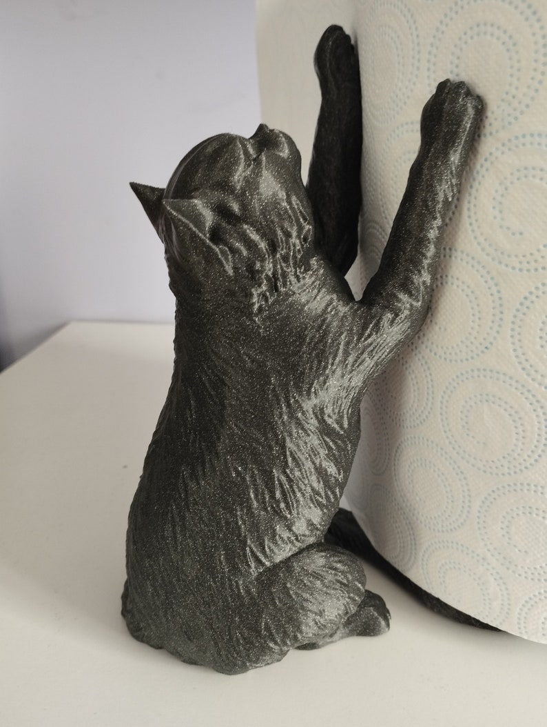 May include: Black cat figurine holding a roll of paper towels. The cat is positioned as if it is climbing the roll of paper towels. The figurine is made of a textured, dark gray material.