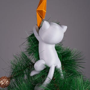 May include: A white cat-shaped Christmas tree topper holding a gold star. The cat is climbing on a green artificial Christmas tree. A gold ornament is visible on a branch. The background is a solid gray.