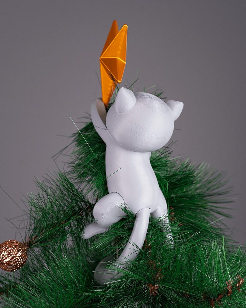 May include: A white cat-shaped Christmas tree topper holding a gold star. The cat is climbing on a green artificial Christmas tree. A gold ornament is visible on a branch. The background is a solid gray.