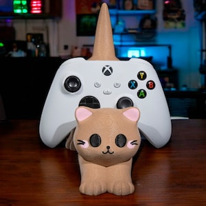May include: A white Xbox controller with a brown cat-shaped stand. The stand has a pink nose and ears and black eyes. The controller is resting on the stand, with the cat's head facing the camera.