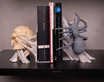 May include: A pair of bookends featuring a skull and a spider. The skull is white and has a textured surface. The spider is gray and has eight legs. The bookends are holding up two books. The book on the left is black with white text that reads "HONORE DE BALZAC". The book on the right is black with white text that reads "CIRCULO E CULTURA".