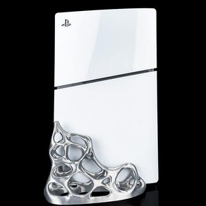 Op de afbeelding: Een witte PlayStation 5 spelcomputer die verticaal staat, met een glanzende afwerking en het PlayStation-logo bovenaan. Een zilverkleurige, metalen standaard met een organische vorm en een poreuze, webachtige structuur is aan de voorzijde bevestigd.