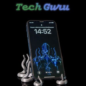 Może przedstawiać: Srebrny stojak na telefon w kształcie ośmiornicy, wydrukowany w 3D, trzymający czarny iPhone z niebieskim tłem ośmiornicy i wyświetlający godzinę 14:52 na ekranie. Telefon znajduje się na czarnej powierzchni.