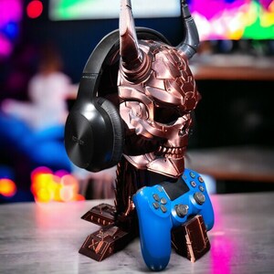May include: A blue gaming controller sits on a metal skull-shaped stand with horns. A black wireless headset is resting on the skull's head.