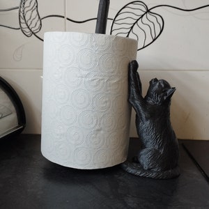 May include: Black cat paper towel holder with a white paper towel roll. The cat is holding onto the roll with its paws and has a playful expression.