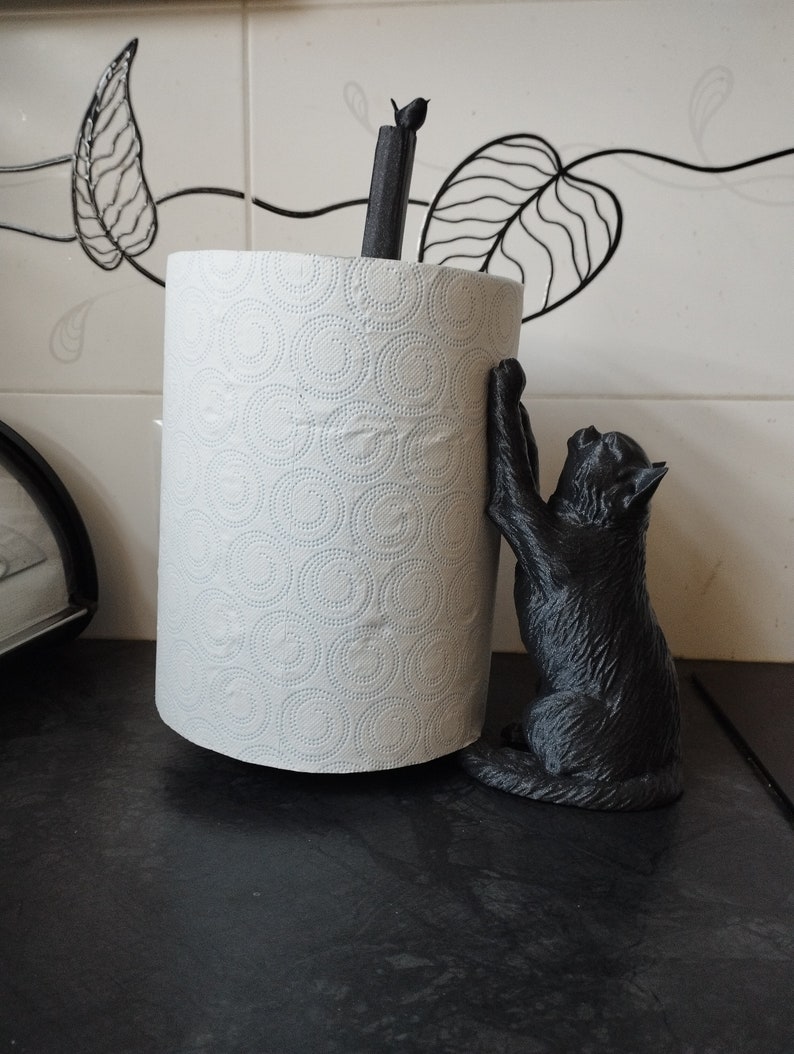 May include: Black cat paper towel holder with a white paper towel roll. The cat is holding onto the roll with its paws and has a playful expression.