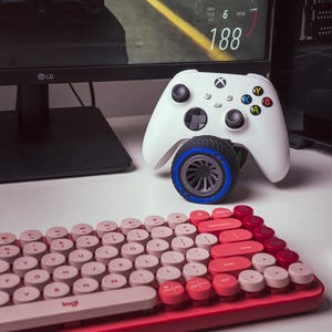 Può includere: Un controller Xbox bianco con una ruota blu e grigia attaccata alla parte inferiore. Il controller è appoggiato su una superficie bianca davanti a un monitor di computer nero. Una tastiera rosa e rossa con tasti rotondi è in primo piano.