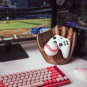 May include: A pink and red retro-style keyboard with round keys, a white baseball, and a white Xbox controller in a brown baseball glove. The glove is sitting on a white desk.