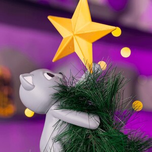 May include: A gray cat figurine holding a green Christmas tree with a yellow star on top. The cat is looking up at the star. The background is a blurred purple with yellow bokeh lights.