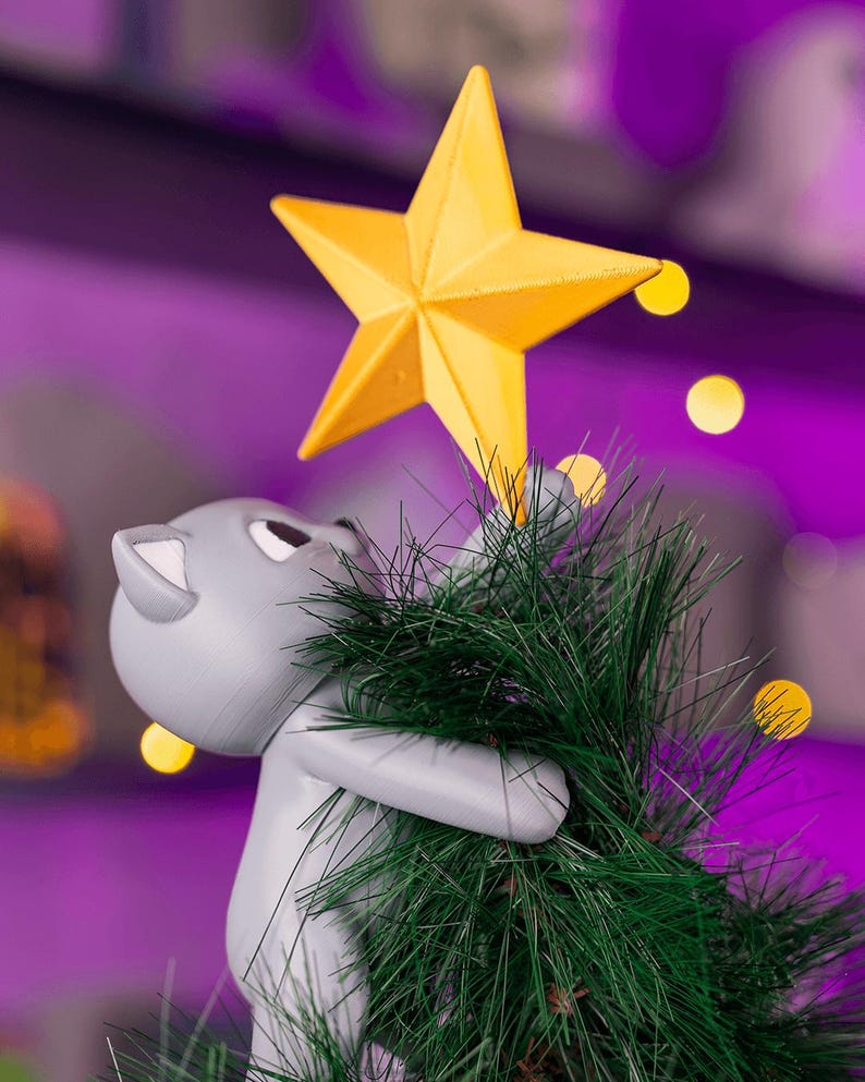 May include: A gray cat figurine holding a green Christmas tree with a yellow star on top. The cat is looking up at the star. The background is a blurred purple with yellow bokeh lights.
