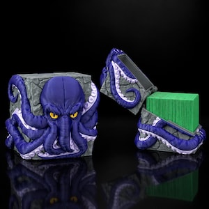 Könnte beinhalten: Eine quadratische Kartenbox mit einem modellierten violetten Oktopus mit gelben Augen, der sich um eine graue, steinstrukturierte Basis windet. Der Deckel ist geöffnet und zeigt einen Stapel grüner Karten, links steht eine zweite Box.