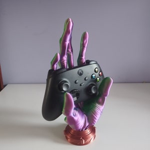 Könnte beinhalten: Ein 3D-gedruckter, handförmiger Ständer für einen Gaming-Controller. Die Hand hat einen Farbverlauf aus Lila, Grün und Bronze und hält einen schwarzen Xbox-Controller. Der Sockel ist bronzefarben. Ein einzigartiges und dekoratives Accessoire für Gamer.