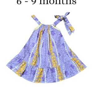 May include: A lavender sleeveless dress with a yellow and white floral pattern and a ruffled hem. The dress features a tie-shoulder strap and the text 6 - 9 months printed above it. The garment has a gathered neckline and a flared silhouette.