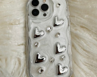 May include: A clear phone case with a white glitter background and silver heart accents. The case has a raised camera bump with three lenses.