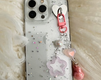 May include: A clear phone case with a silver glitter design and a pink heart charm attached to a chain with pink and white beads. The charm features a star, a heart, and a small pink bunny.
