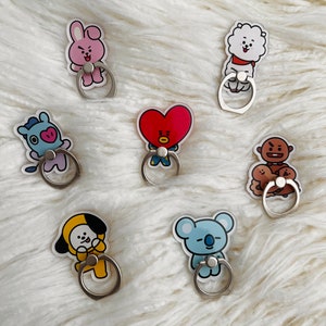 May include: Seven different phone ring holders featuring cartoon characters from the popular K-pop group BTS. The characters are all in different poses and expressions. The holders are made of clear plastic with a silver metal ring.