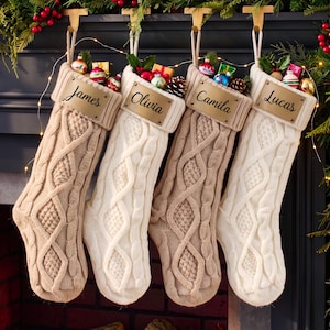 May include: Four cable-knit Christmas stockings in beige and white, each featuring a rectangular leather-like tag with the names James, Olivia, Camila, and Lucas engraved in black script. The stockings are filled with small colorful ornaments.