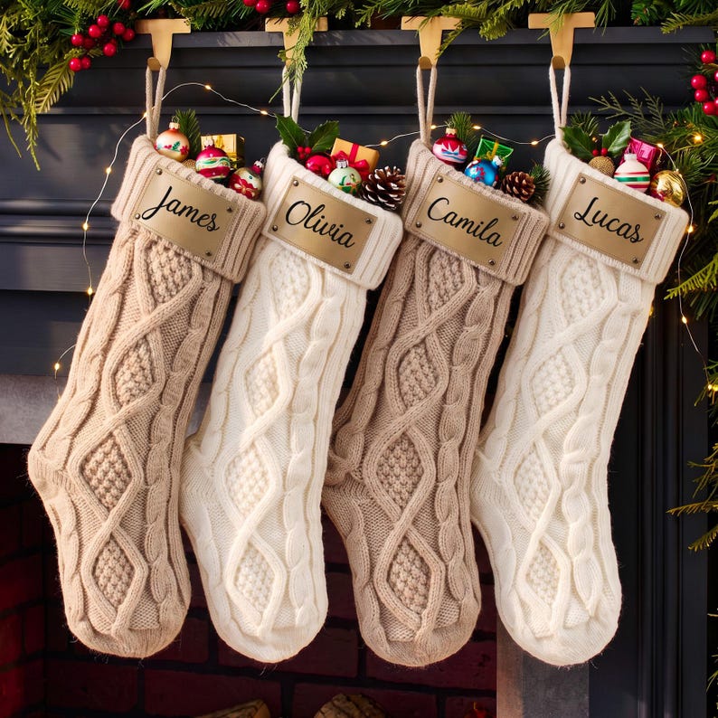 May include: Four cable-knit Christmas stockings in beige and white, each featuring a rectangular leather-like tag with the names James, Olivia, Camila, and Lucas engraved in black script. The stockings are filled with small colorful ornaments.
