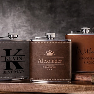 May include: Three personalised brown leather-wrapped stainless steel hip flasks with engraved names and titles. The flasks are on a wooden surface.