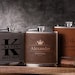 May include: Three personalised brown leather-wrapped stainless steel hip flasks with engraved names and titles. The flasks are on a wooden surface.