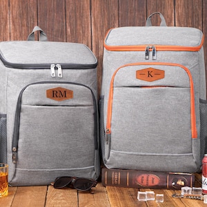 May include: Two gray insulated backpacks with personalized leather patches. One backpack has orange trim and the other has black trim. Both have a top handle and a front pocket. Sunglasses and a glass of amber liquid are in front of the backpacks.
